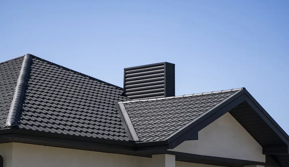A close-up view of a modern house roof with dark gray, overlapping roof tiles and a rectangular vent structure against a clear blue sky.