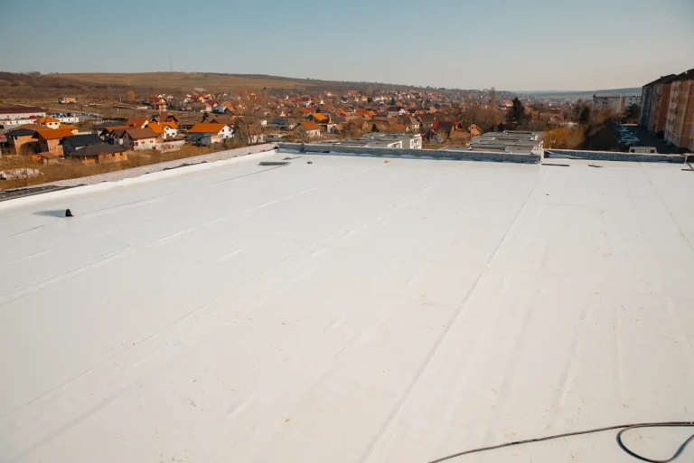 A large, flat white roof with a smooth surface overlooks a suburban area with houses, buildings, and rolling hills in the background under a clear sky.