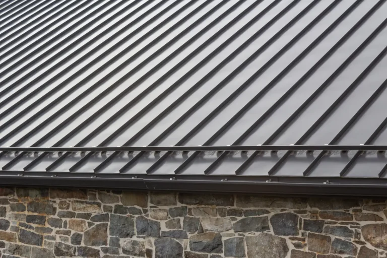Close-up view of a sloped metal roof with standing seams on top of a stone wall, showing the roofs shiny, corrugated panels and the irregular pattern of stones in the wall below.