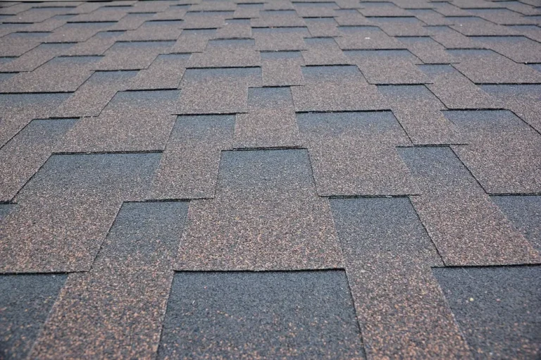 Close-up view of a roof covered with overlapping, rectangular asphalt shingles in shades of black and brown, arranged in a staggered pattern.