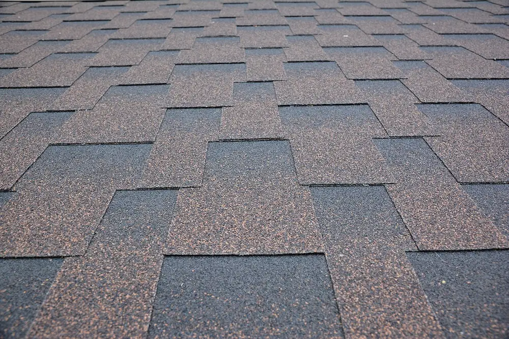 Close-up view of a roof covered with overlapping, rectangular asphalt shingles in shades of black and brown, arranged in a staggered pattern.