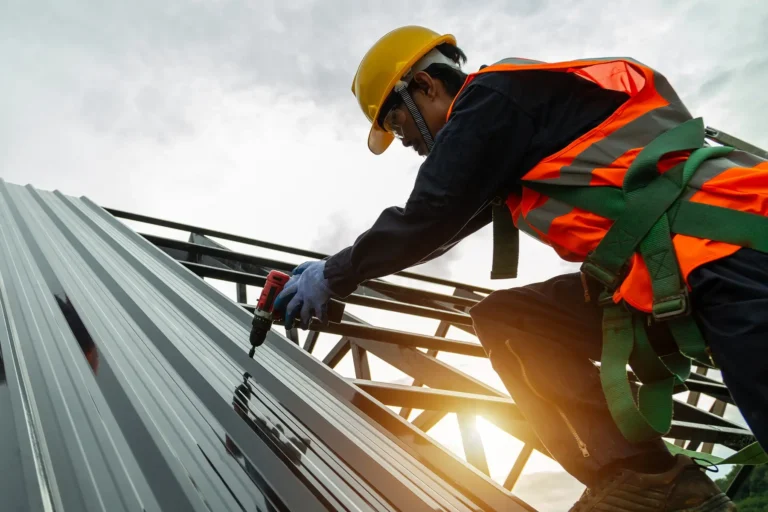 A construction worker wearing a hard hat, safety harness, gloves, and orange vest uses a power drill to install metal roofing sheets on a building frame under a cloudy sky.