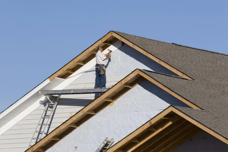 A construction worker stands on scaffolding, installing siding on the exterior of a house under construction, with exposed wooden beams and a clear blue sky in the background.