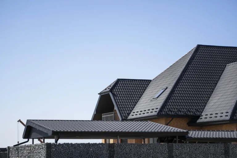 Modern house with dark gray, sloped metal roofs and a skylight, partially visible behind a stone fence, set against a clear blue sky.