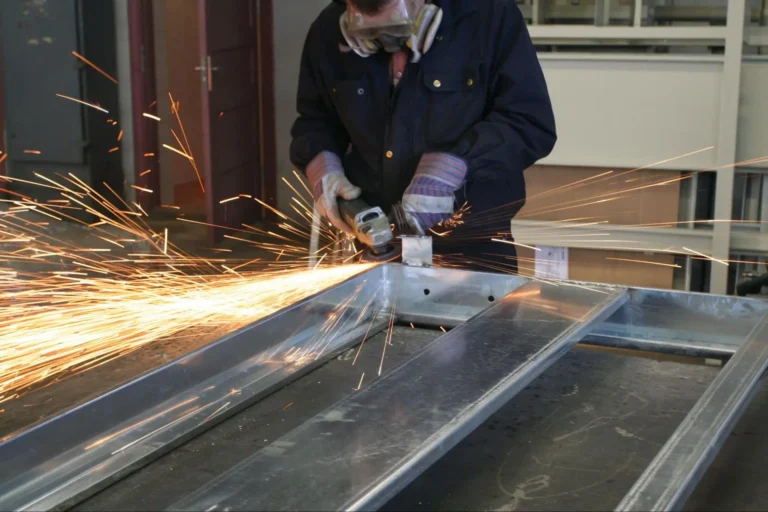 A person wearing protective gear uses an angle grinder on a metal frame, producing bright sparks in a workshop setting.