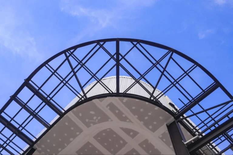 A low-angle view of a metal arch structure with a geometric pattern, partially covering a dome-like building, set against a clear blue sky.