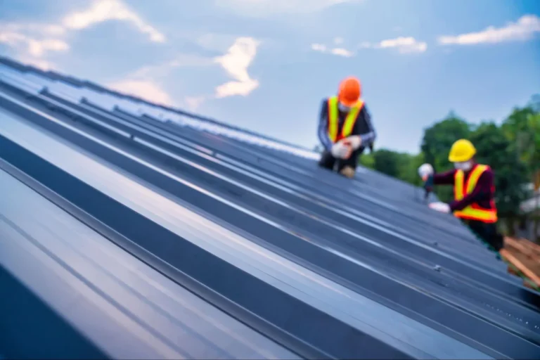 Two construction workers in safety vests and helmets work on a sloped metal roof, with the focus on the roof panels and a blurred background of trees and sky.