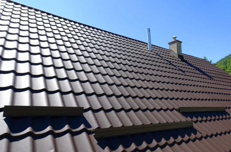 A close-up view of a sloped brown metal tile roof with chimney and vent pipes under a clear blue sky. The roof tiles have a wavy pattern and are evenly aligned.