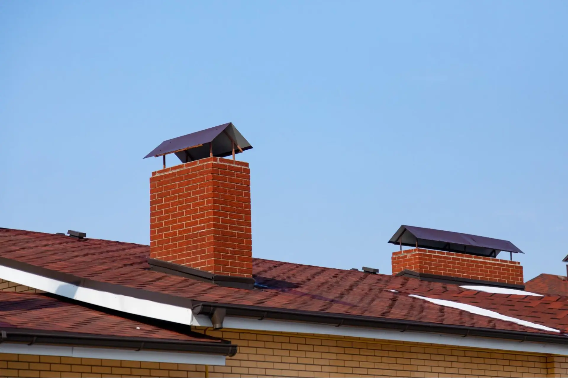 Two brick chimneys with metal caps are mounted on the sloped, reddish-brown roof of a house, set against a clear blue sky. The house has yellow brick walls and white trim.