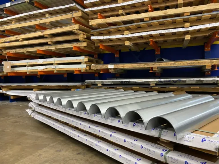 Stacks of corrugated metal roofing sheets are stored on wooden racks in a warehouse with other construction materials visible on shelves in the background.