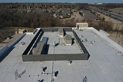 Aerial view of a large flat commercial building roof with several HVAC units enclosed by a fence, surrounded by other buildings, trees, and a nearby road.