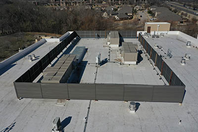Aerial view of a flat, white commercial rooftop with HVAC units enclosed by tall gray metal screens, surrounded by other rooftop equipment and neighboring buildings in the background.