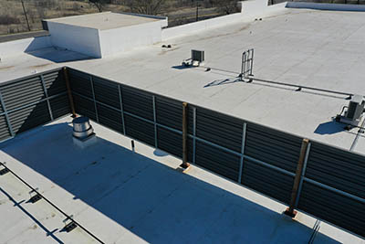 Aerial view of a flat commercial building roof with HVAC units, a large metal louvered screen, and rooftop vents under clear daylight.