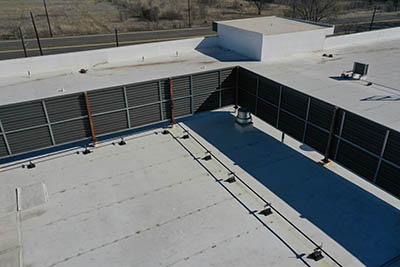 A flat white rooftop with a metal louvered wall and some rooftop equipment, viewed from above. A road and dry grassy land are visible in the background.