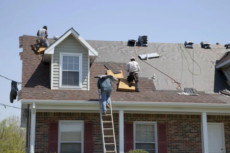 Three workers are installing new shingles on the roof of a brick house. One is on a ladder, while the other two are on the roof, working with tools and materials under a clear blue sky.
