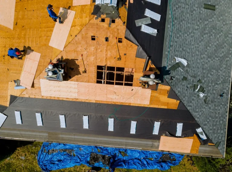 Aerial view of workers installing plywood panels and roofing materials on a house roof under renovation, with tools and equipment visible and a blue tarp spread on the ground below.