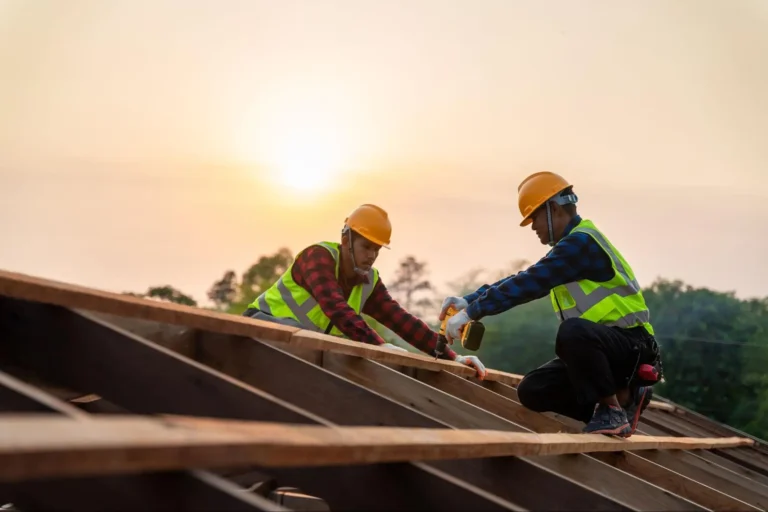 Two construction workers in safety vests and helmets work on a wooden roof structure at sunset, using tools to secure beams, with trees and a bright sky in the background.