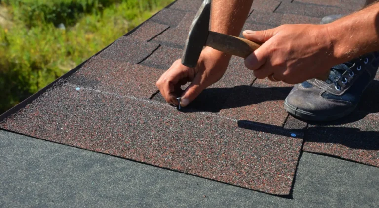 A person uses a hammer to drive a nail into asphalt shingles while installing roofing on a house. The persons hands and part of a shoe are visible on the roof.
