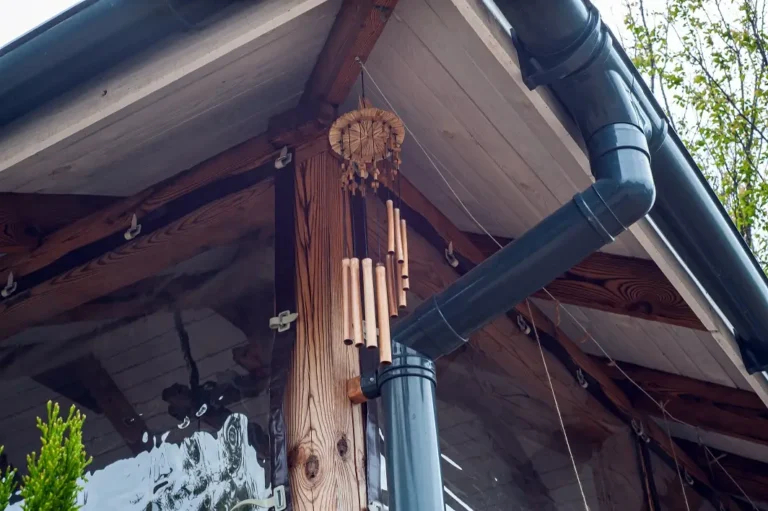 A wooden wind chime hangs from the corner of a wooden roof near a black rain gutter on a house, with a clear plastic panel enclosing the porch area.