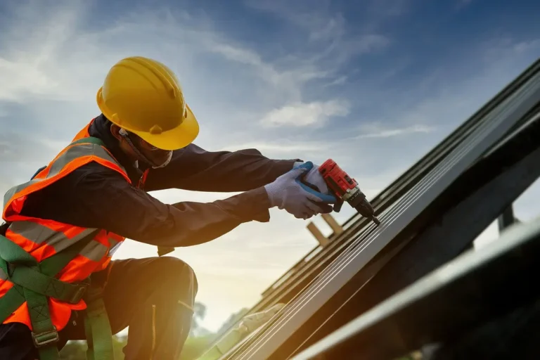 A construction worker wearing a yellow hard hat, safety vest, gloves, and safety glasses uses a power drill on a metal structure while working on a sloped roof at sunset.