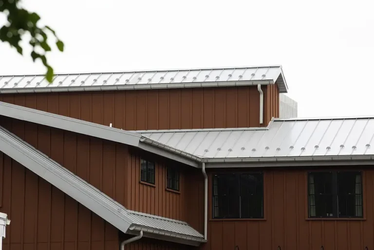 A close-up view of a modern building with brown vertical siding and multiple angled, silver metal roofs under an overcast sky.