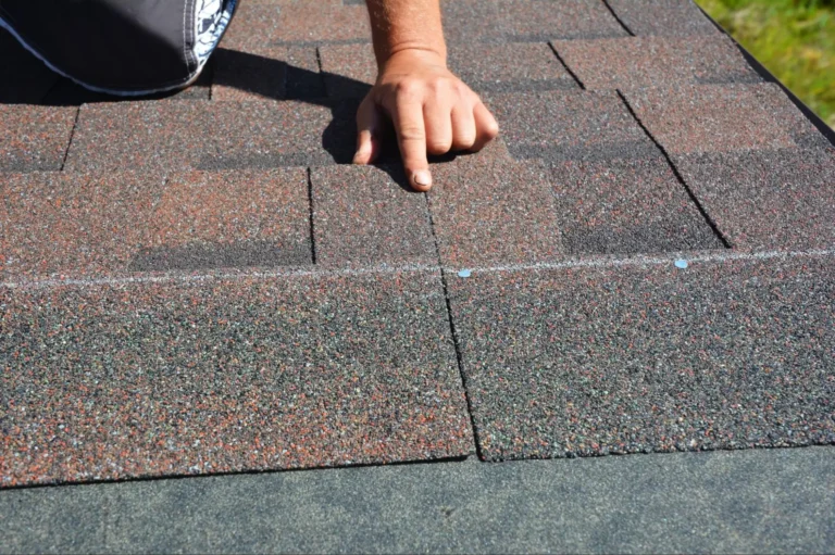 A person’s hand is pressing down on newly installed asphalt roof shingles, aligning the shingles along a straight chalk line on a rooftop.