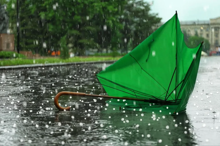 A green umbrella lying upside down on a wet street during heavy rain, with raindrops splashing on the ground and blurred trees in the background.