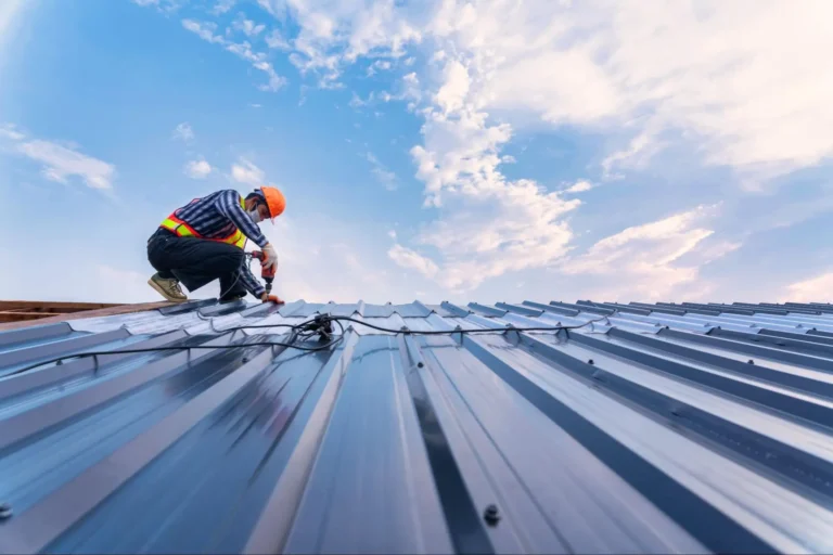 A worker in a safety vest and helmet installs or repairs electrical wiring on a metal rooftop under a partly cloudy sky.