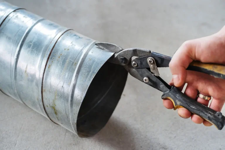 A hand demonstrates how to cut sheet metal using metal snips, slicing through a cylindrical duct placed on a gray surface.