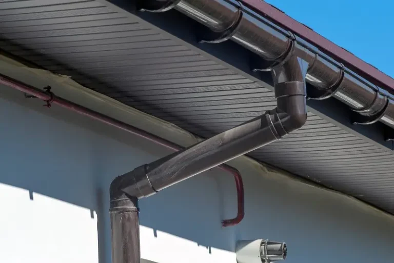 Black rain gutter and downspout attached to the edge of a house roof, with blue sky in the background. The gutter system is angled and runs along the roofline and down the building’s exterior wall.