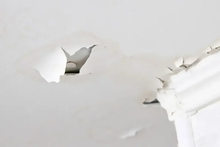 A close-up of a white ceiling with peeling paint and water damage, showing cracks and a hole exposing the underlying material.