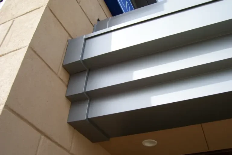 Close-up view of an architectural detail featuring a beige stone wall and a modern, metallic overhang with horizontal, rectangular gray panels and a recessed light underneath.
