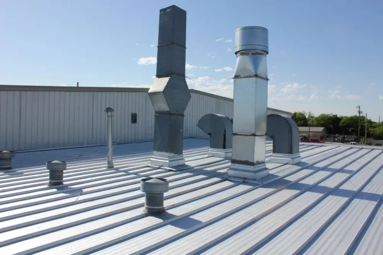 A metal rooftop with several ventilation pipes and ducts of varying shapes and sizes, with a light industrial building and clear blue sky in the background.
