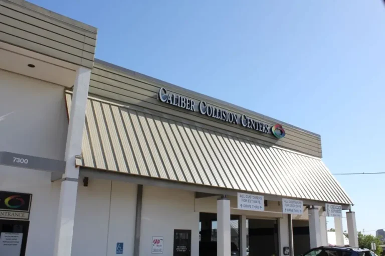 The exterior of a building with a sign that reads Caliber Collision Center. The building has a slanted metal roof, several columns, and visible signs near the entrance under a clear blue sky.