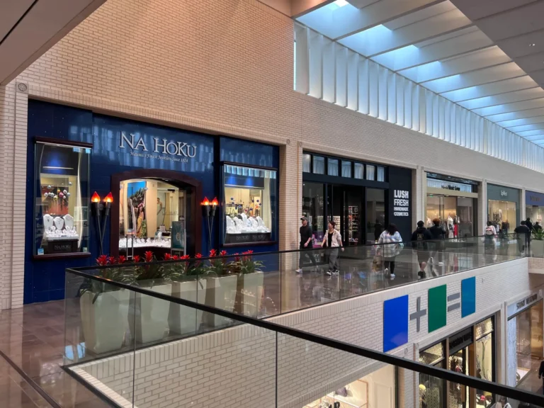 A view of an indoor shopping mall’s upper level, showing several store fronts including Na Hoku jewelry, with shoppers walking and sitting on benches along the corridor. The mall has large skylights and modern decor.