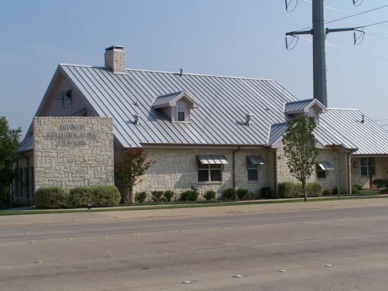 A stone building with a metal roof, small dormer windows, and a chimney sits beside a wide street. Large power lines and neatly trimmed shrubs are visible in front of the building.