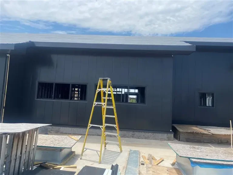 A yellow ladder stands in front of a dark gray building under construction, with open window frames and construction materials scattered on the ground. The sky is partly cloudy above.