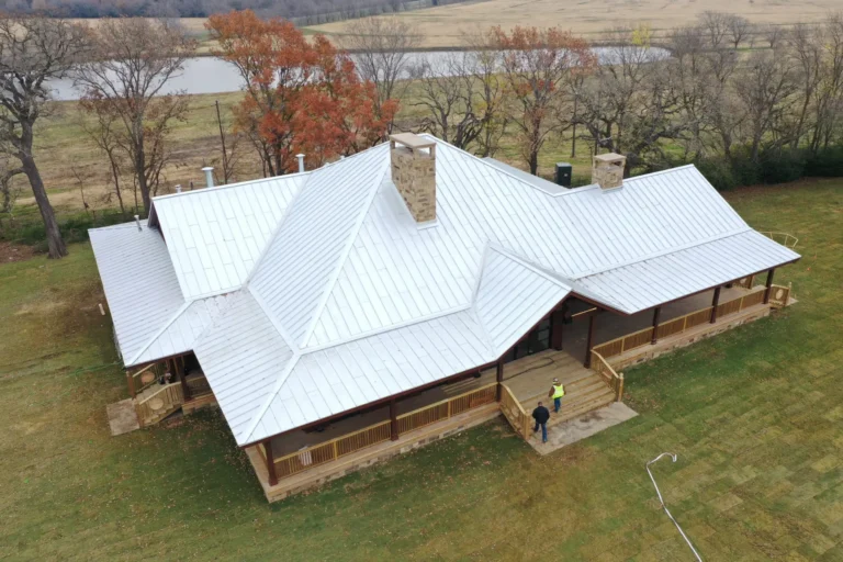 Aerial view of a large house with a white metal roof, wraparound porch, and stone chimneys, surrounded by grassy lawn, trees with fall foliage, and a pond in the background. Two people stand at the front steps.