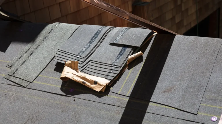 A stack of asphalt roofing shingles and packaging material rests on a partially shingled roof under sunlight, with wood siding visible in the background.