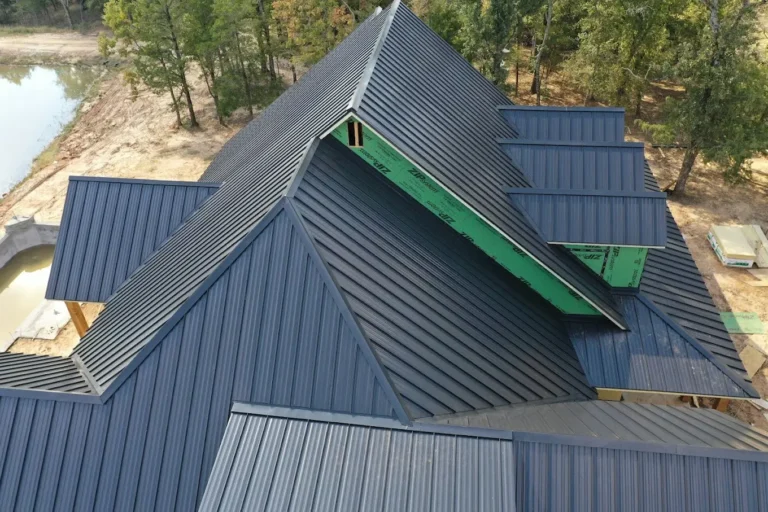 Aerial view of a modern house with a complex, angular metal roof. The roof features dark panels and geometric shapes. Surrounding the house are trees, a small body of water, and a partially constructed area.