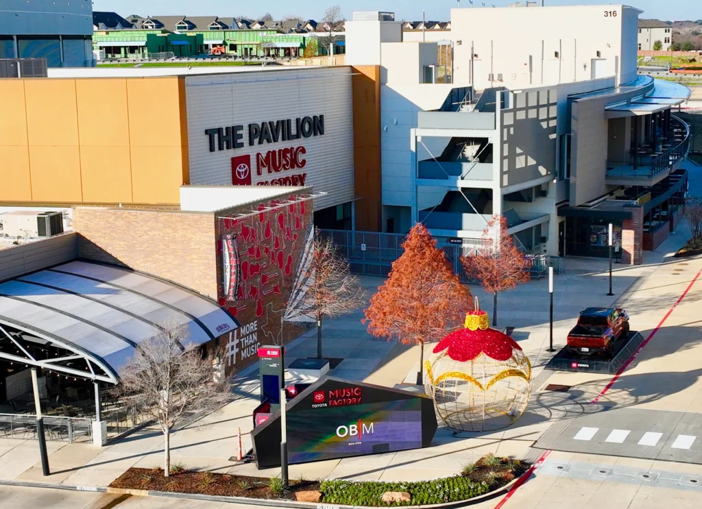 custom flashing & trim An outdoor view of The Pavilion at Toyota Music Factory, showcasing modern buildings with sleek commercial sheet metal accents, a parking garage, trees with autumn leaves, and a large decorative ornament on the sidewalk.