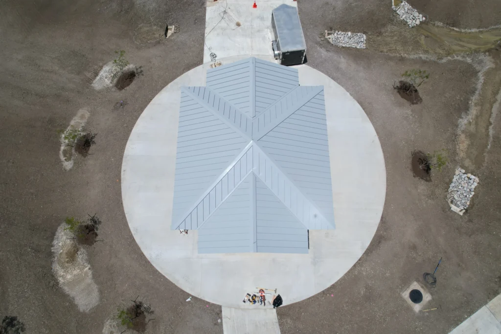 standing seam metal roofing Aerial view of a small, gray, metal-roofed building centered on a circular concrete pad, surrounded by dry, brown ground with sparse vegetation and a trailer parked nearby.
