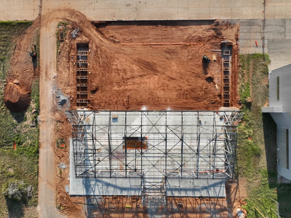 standing seam roofers Aerial view of a construction site showing a rectangular concrete foundation with a steel framework. Surrounding the structure is red dirt, equipment, and workers, with a dirt road curving along one side.