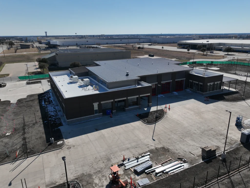 architectural metal soffit systems Aerial view of a newly constructed, modern, single-story building with a metal roof, surrounded by paved roads and some construction materials; industrial buildings and open land are visible in the background.