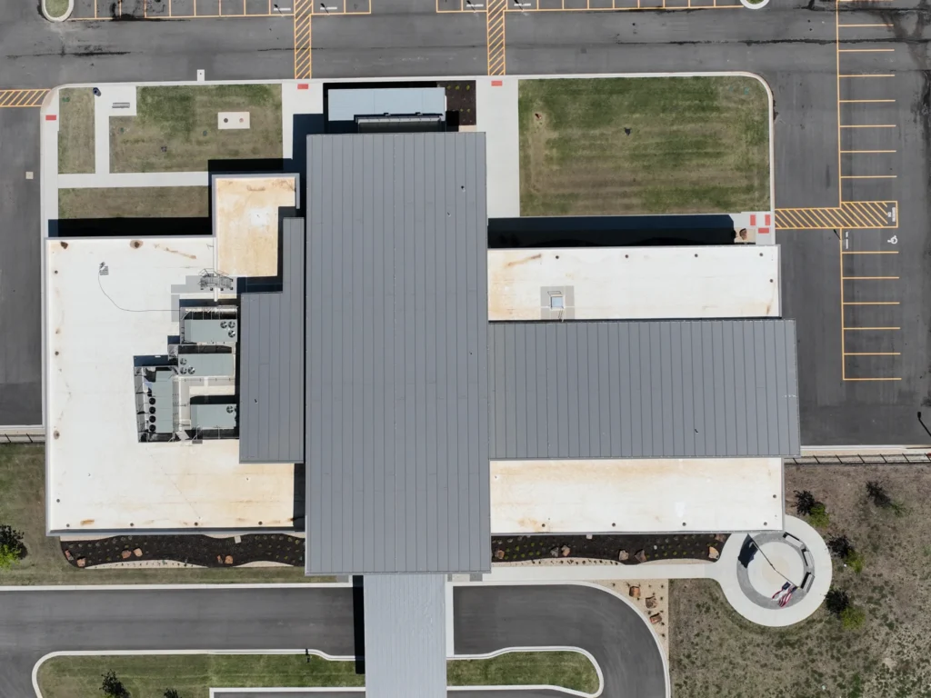 Aerial view of a modern building with a gray roof, surrounded by parking lots, green lawns, and roads. The layout of the structure forms a T-shape, with clearly marked parking spaces and sidewalks.