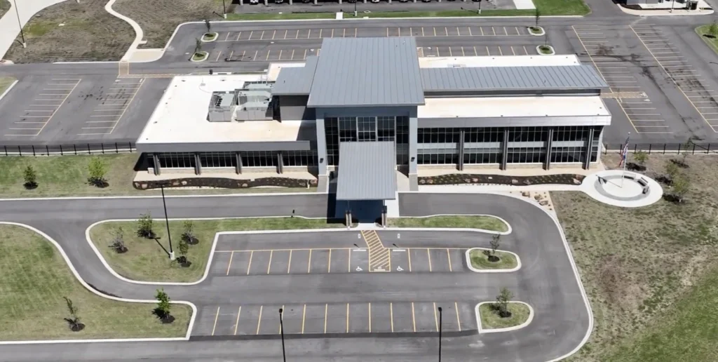 Aerial view of a modern two-story building with a metal roof, large windows, and an entrance canopy. The building is surrounded by empty parking lots and landscaped areas.