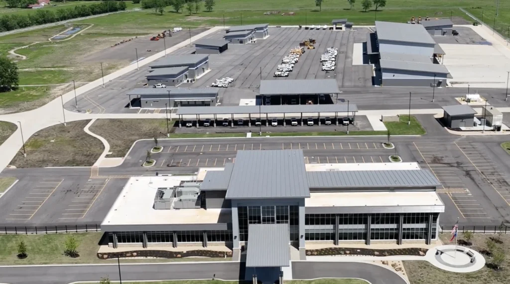 Aerial view of a large modern industrial or office complex with multiple gray-roofed buildings, parking lots with vehicles, and landscaped green areas surrounding the property.
