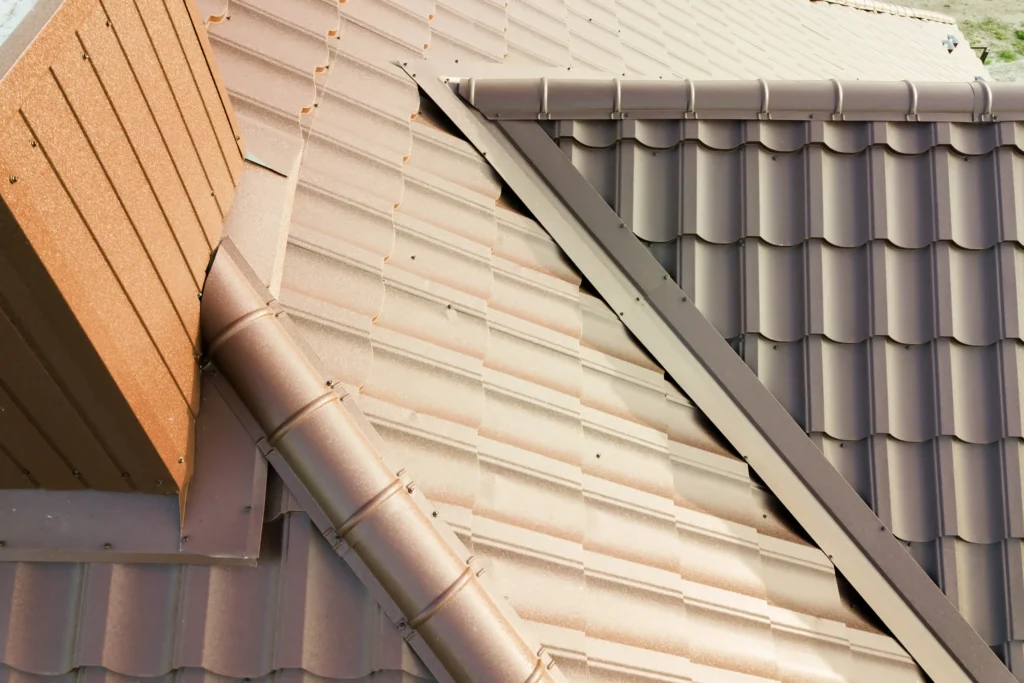 metal gutters Close-up view of a house roof with overlapping brown metal tiles and joints, showing different roof angles and ridge caps. The roofing appears clean and newly installed.