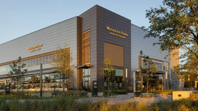 A modern, two-story building with large glass windows and dark paneling houses Mendocino Farms, a sandwich market. The area is landscaped with young trees and grasses under a clear sky.