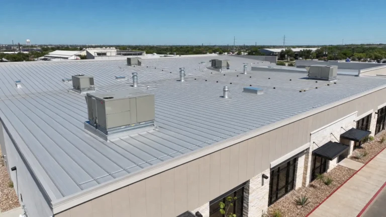 Aerial view of a commercial building with a large, flat metal roof featuring multiple HVAC units and ventilation pipes under a clear blue sky.